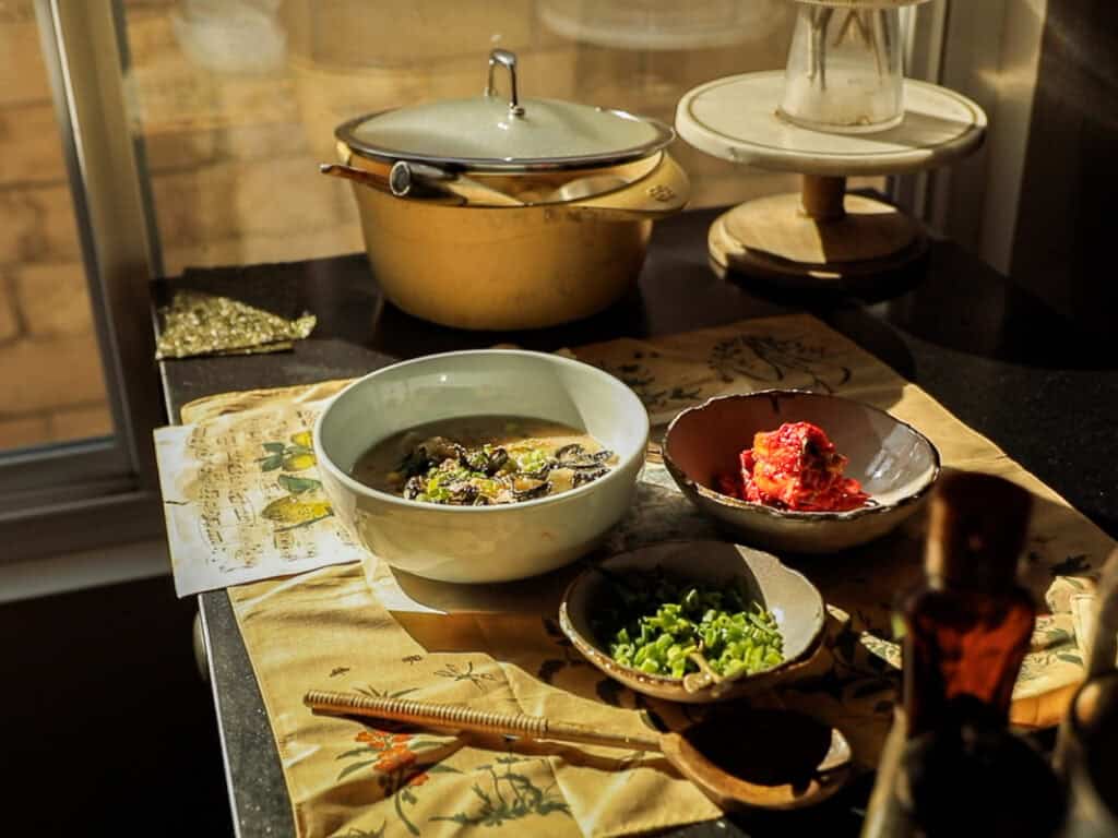 A sunlit table with a bowl of soup and a red dish, surrounded by a small bowl of chopped green onions, a metallic pot with a lid, and a wooden utensil. A yellow printed cloth covers the table surface.